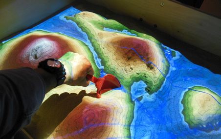 <p>Surplombé d'une caméra 3D et d'un projecteur vidéo, le bac à sable interactif utilise la réalité augmentée pour enseigner les principes de la géologie.</p>