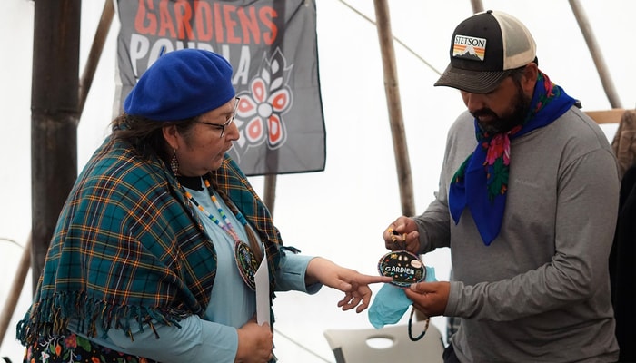 <p>Durant la graduation dans le grand shaputuan au site du Mushuau-nipi. L’aînée innue de Mashteuiatsh, Doris Bossum, en compagnie de son neveu Stacy Bossum, étudiant ayant reçu la certification de gardien.nes de territoire. Celui-ci travaille au soutien d’initiatives en développement culturel pour les Pekuakamiulnuatsh, les Innus du Lac-Saint-Jean. Doris Bossum a confectionné le médaillon à la main en ajoutant des motifs inspirés du travail et des implications de son neveu.</p>