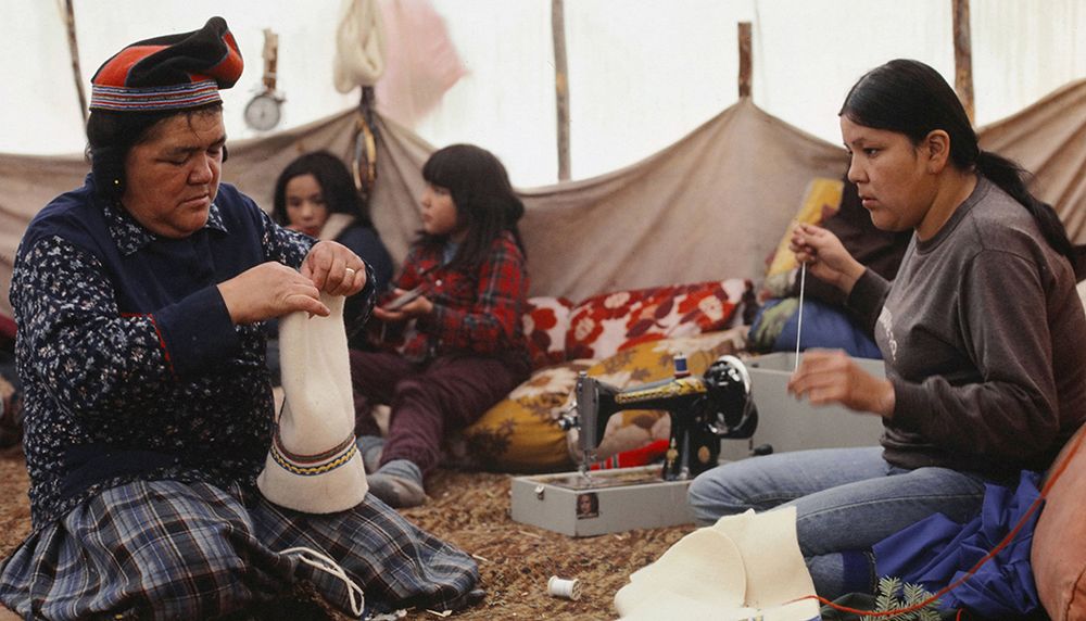 Novembre 1982. À l’intérieur de la tente, au lieu de campement de Nishk-pineiauit, Marie-Paule Mark aide sa mère Hélène dans la confection de mocassins d’hiver à partir d’un type de toile qui sert également à la confection de la tente, des vêtements, des sacs de portage et des canots. Les enfants Anikuni et Suzie sont à l’arrière.