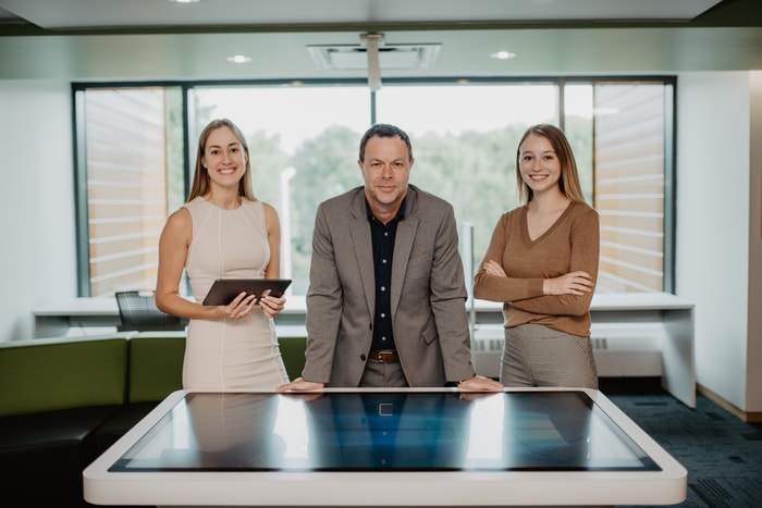 <p>À l’entrée des bureaux de l’UMR-SU. Gabrielle Teyssier-Roberge, Sébastien Tremblay et Sarah Savage derrière une table numérique tactile interactive, l’un des outils technologiques à la disposition de l’UMR pour des démonstrations ou des présentations.</p>