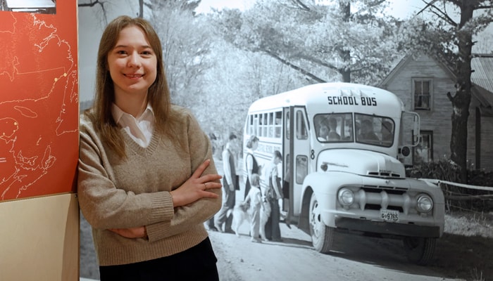 La commissaire Geneviève Boisvert devant l'une des très grandes photographies d'époque en noir et blanc qui enrichissent l'exposition. Celle-ci est l'œuvre de Joseph Guibord en 1951. On y voit des élèves prenant l'autobus scolaire en direction de l'école de Knowlton, dans la région des Cantons-de-l'Est, au Québec. Le passage de la carriole tirée par des chevaux à l'autobus scolaire motorisé change radicalement la réalité des commissions scolaires des régions du Québec.