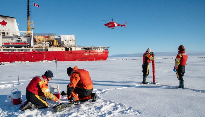 Des scientifiques procèdent à une collecte de données sur la banquise, avec l'hélicoptère et le brise-glace Amundsen en arrière-plan.