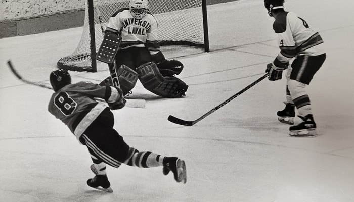 Hiver 1983: le gardien de but du Rouge et Or, Paul Maynard, stoppe un tir d'un joueur de l'Université du Québec à Chicoutimi. Serge Turcotte (no 4) attend le retour. Division des archives de l'Université Laval, fonds Service des activités sportives.
