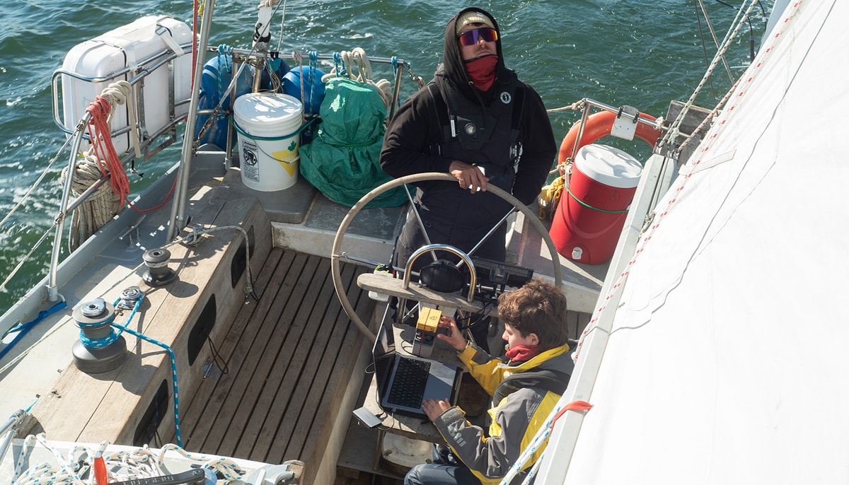 Poste de pilotage du voilier avec le skipper Bastian Raulier et une étudiante utilisant un outil d'imagerie du plancton pour observer les organismes récoltés dans le filet à phytoplancton